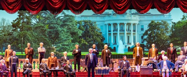 Hillary Clinton (center, in a man's business suit) leads the Hall of Presidents. Photo by Steve Wise [CC BY-SA 2.0] via Flickr.