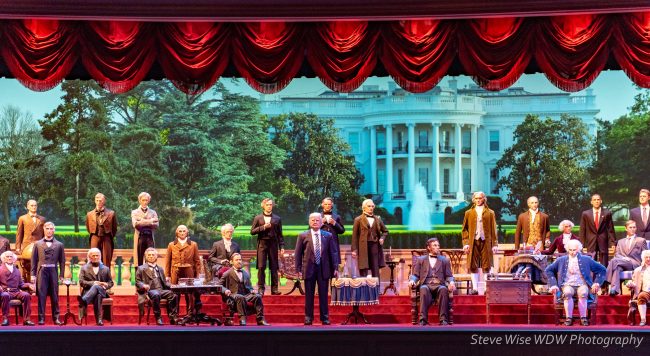 Hillary Clinton (center, in a man's business suit) leads the Hall of Presidents. Photo by Steve Wise [CC BY-SA 2.0] via Flickr.